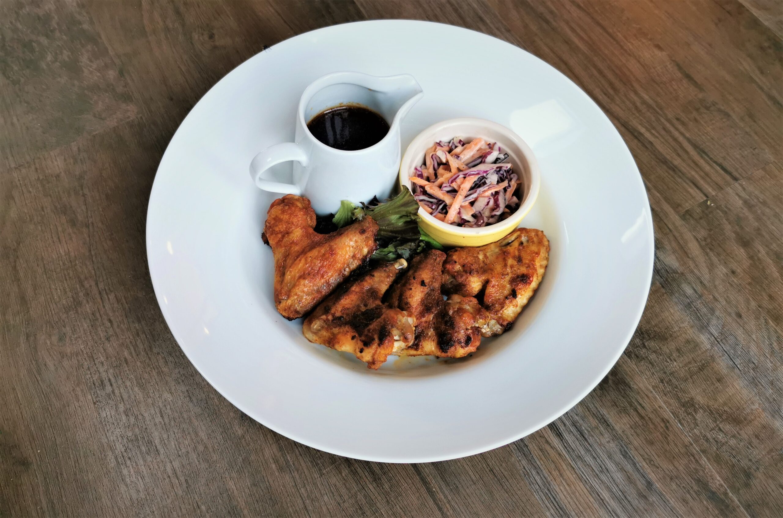a wooden surface with a white bowl upon which is four golden chicken wings, salad, a ramekin of coleslaw and a jug with sauce