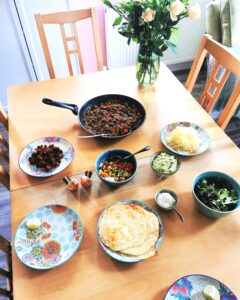 A wooden table laid with colourful plates, a pan of mexican meat, guacamole, creme fraiche, grated cheese, salsa, roasted cauliflower and salad. There is also a vase with flowers.