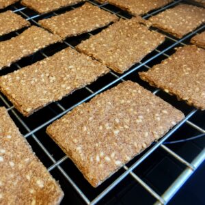 A close up of oblong low carb crackers with sesame seeds sitting on a wire rack over a black background